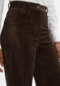 Brązowe spodnie z korduroyu o poziomej struktury, wysoki stan, boczne kieszenie oraz metalowe zapięcie, z paskami na pasek w pasie.