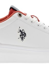 Zapatilla de cuero blanca con acentos de malla roja, cordones planos y un logo de jugador de polo negro. Presenta suela de goma texturizada y detalles de ojales metálicos.