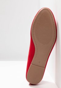 Chaussure plate en textile rouge appuyée contre une surface blanche. La semelle est beige clair avec un motif strié pour une meilleure traction.