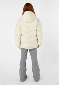 Cremfarbene Pufferjacke mit Kapuze, die ein gestepptes Design und elastische Bündchen aufweist. Kombiniert mit grauen, figurbetonten Hosen. Rückansicht des Outfits.