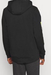 Persona che indossa una felpa nera con cappuccio e una grafica giallo neon sul braccio sinistro, abbinata a pantaloni grigio scuro, vista da dietro.