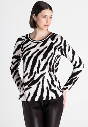Femme aux cheveux courts et foncés portant un haut à manches longues imprimé zèbre noir et blanc et un pantalon noir, regardant sur le côté avec la main sur la hanche.