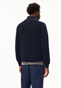 Mann mit kurzen lockigen Haaren, der einen dunkelblauen gerippten Strickpullover und blaue Denim-Cargohosen trägt, mit dem Rücken zu sehen vor einem weißen Hintergrund.