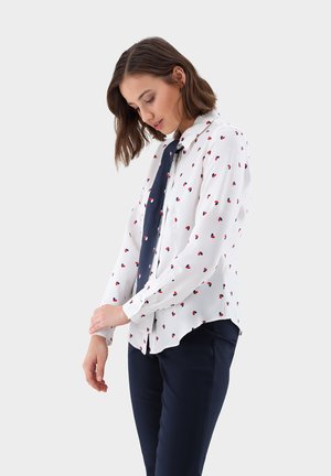 Weiße Bluse mit rotem Herzenmuster und langen Ärmeln, begleitet von einer dunkelblauen Krawatte. Kombiniert mit marineblauen Hosen, die einen lässigen Schnitt zeigen.
