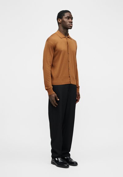 Άνδρας που στέκεται φορώντας καφέ πλεκτή ζακέτα με κουμπιά, μαύρο παντελόνι και μαύρα παπούτσια ντυσίματος μπροστά σε απλό λευκό φόντο.