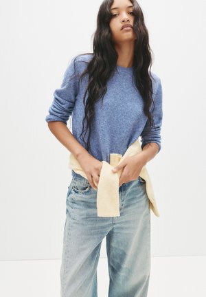 Blauer Wollmischpullover mit gerippten Bündchen, kombiniert mit hellblauen Jeans. Ein gelber Strickpullover ist um die Taille gebunden.