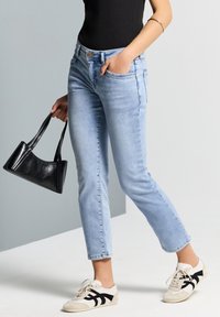 Jeans de mezclilla azul claro con corte slim, bolsillos laterales y una pierna ligeramente entallada, combinados con un bolso negro y zapatillas informales.
