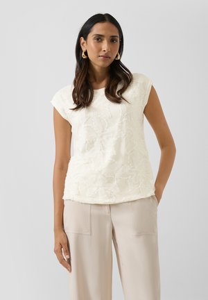 Frau mit langen dunklen Haaren, die goldene Creolen, ein weißes kurzärmeliges Oberteil mit floralem Muster und eine beige Hose mit weitem Bein trägt.