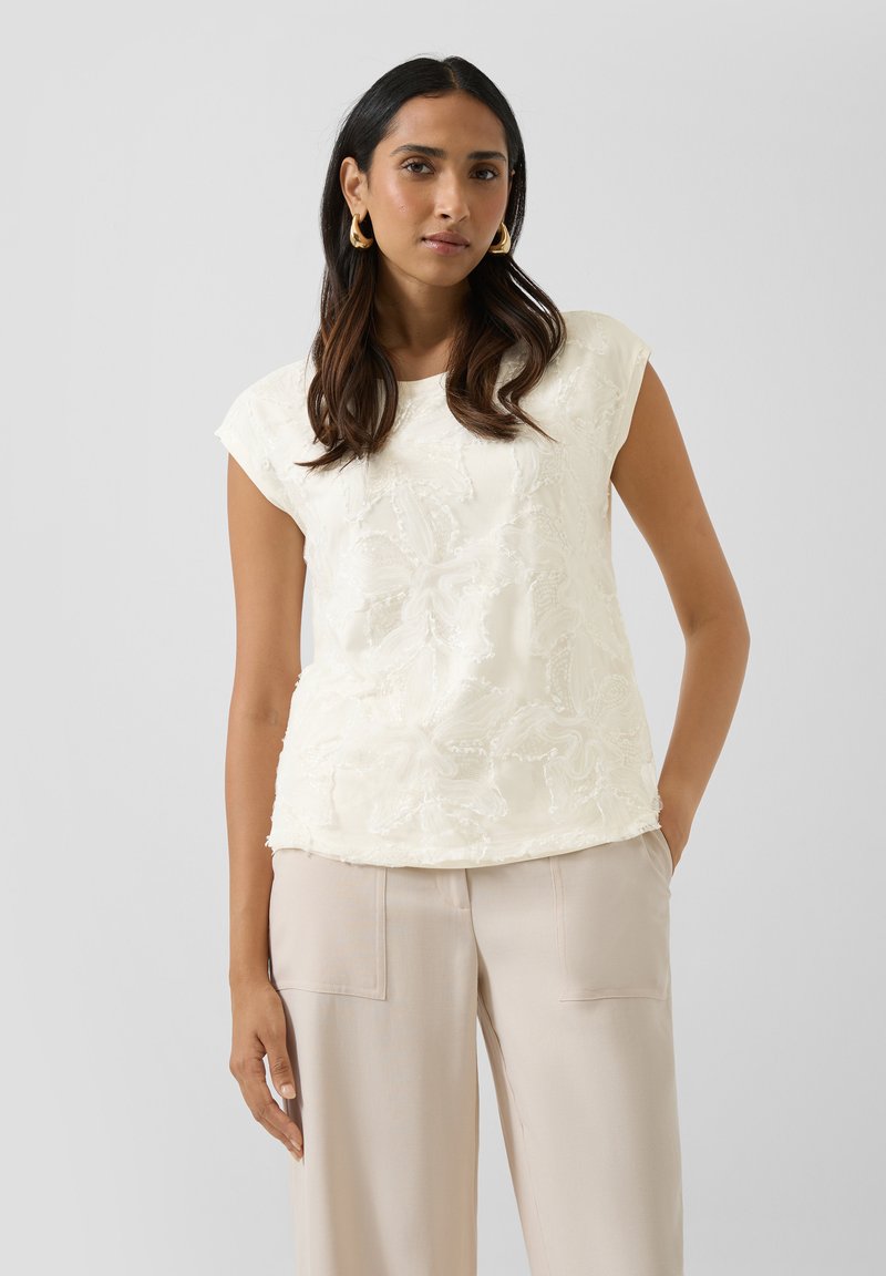 Vrouw met lang donker haar, gouden oorbellen in hoefijzervorm, een wit topje met korte mouwen en bloemenstructuur, en beige wijduitlopende broek.