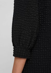 Svart texturerad klänning med ruta-mönster och puffärmar. Tyget ser skrynkligt ut, vilket ger djup och dimension.