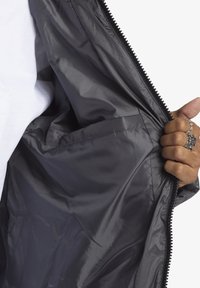 Grå jacka på insidan med en slät, glänsande textur och dragkedjestängning; inkluderar en ficka på vänster sida.