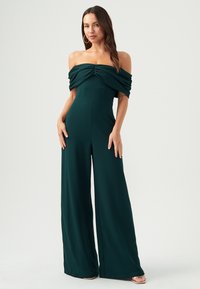 Jumpsuit bez ramínek v tmavě zelené barvě se nařaseným korpusem, širokými nohavicemi a hladkým materiálem. Má těsně padnoucí pas a jemné švy.