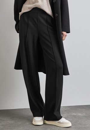 Person trägt schwarze Hose mit weitem Bein, beige Pullover, schwarzen langen Mantel und weiße Sneaker und steht auf grauem Boden vor einer schlichten Wand.