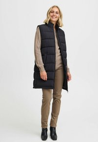 Gilet noir rembourré avec col montant, poches latérales et design matelassé, porté sur un pull beige et un pantalon beige, complété par des bottes noires.
