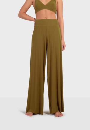 Vrouw draagt olijfgroene wijde pantalon en bijpassende cropped mouwloze top, staat blootsvoets op een effen witte achtergrond.