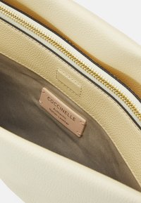 Inredning av en krämfärgad läderväska med ett guldfärgat zipsystem, med en etikett som läser "Coccinelle Autentic Italiensk Läder" mot en neutral tygbakgrund.