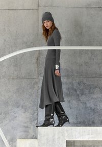 Nő szürke kötött ruhában, hozzáillő sapkával és fekete térdig érő csizmával, egy betonlépcsőn áll, háttérben egy szürke, texturált fallal.