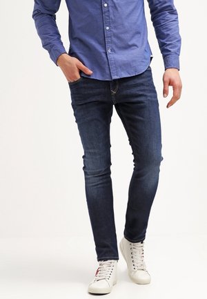Mann trägt dunkelblaue Slim-Fit-Jeans, blaues Hemd mit Knöpfen und weiße High-Top-Sneaker, steht mit einer Hand in der Tasche.