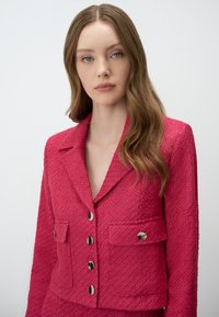 Blazer texturé fuchsia avec un col cranté, des boutons en métal argenté et deux poches avant. Le tissu présente un motif tissé et une coupe cintrée.