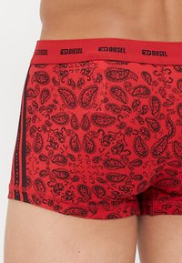 Gros plan sur un boxer rouge avec un motif paisley noir et un logo Diesel noir sur la ceinture élastique porté par une personne.