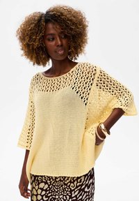 Luźny żółty sweter z dzianiny o szerokim dekolcie, charakteryzujący się kontrastującym wzorem otwartych splotów i krótkimi rękawami. Zestawiony z wzorzystą spódnicą.