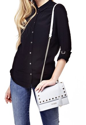 Svetle šedá kožená kabelka s zlatými hroty, řetízkem na rameno a klopovým uzavřením, s vyraženým názvem značky "GUESS" na přední straně.