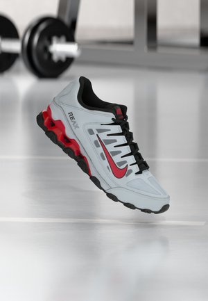 Scarpa sportiva Nike Reax bianca e rossa con dettagli neri, che fluttua sopra un pavimento da palestra con manubri sfocati sullo sfondo.