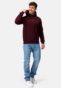 Hoodie bordeaux avec cordons de serrage réglables et logo doré, associé à un jean bleu clair et à des baskets blanches. Texture lisse, coupe décontractée.