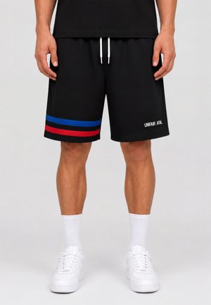 Mand iført sorte shorts med røde og blå striber på venstre ben og hvid tekst på højre ben, kombineret med hvide sokker og hvide sneakers.