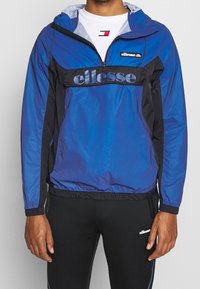 Blue and black windbreaker jacket with a quarter-zip closure, adjustable hood, and a large "ellesse" logo on the front. Made of lightweight material.