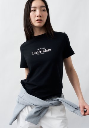 Fiatal nő fekete Calvin Klein pólóban fehér felirattal, fehér nadrágban, és világosszürke pulóverrel, amelyet a derekára kötött.