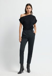 Femme portant un top noir à une épaule, un pantalon noir ajusté et des bottines à talons noires, se tenant devant un fond blanc uni.