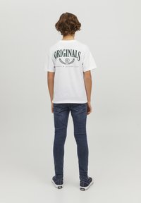 Camiseta de algodón blanca con "ORIGINALS" y ramas de laurel impresas en verde en la parte de atrás, combinada con jeans ajustados de color azul oscuro y zapatillas navy.