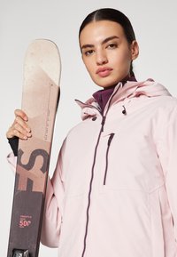 Šviesiai rožinė slidinėjimo striukė su aukštu apykakle ir purpurinėmis akcentais, derinama su beige ir juodomis slidėmis su tekstiniu dizainu šone.
