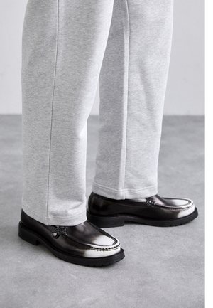 Black loafers with a smooth texture and light gray accent along the edge, paired with gray sweatpants. Low-profile rubber sole.