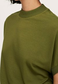 Oliwkowa bluzka z krótkim rękawem, z prążkowanym okrągłym dekoltem, wykonana z gładkiego materiału i o luźnym kroju. Brak widocznych wzorów lub akcentów.