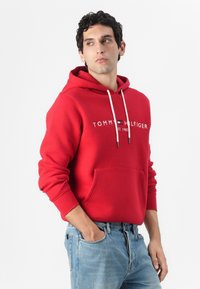 Rød hettegenser laget av bomullsblanding, med en frontlomme, snøring i hetta og hvit merkevarelogo over brystet. Kombinert med blå denimjeans.