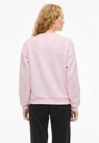 Svetlo roza sweatshirt z okroglim vratom in dolgimi rokavi. Ima sproščen kroj in mehko, gladko teksturo. V kombinaciji z črnimi hlačami.