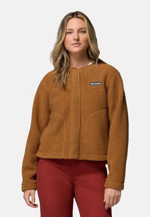 Young woman with long blonde hair wearing a brown fleece jacket and red pants, standing against a plain light background.
