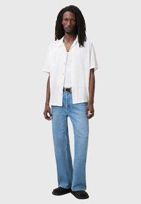 Camicia bianca a maniche corte con texture, jeans larghi blu chiaro, scarpe nere, collana d'argento, in piedi su uno sfondo neutro.