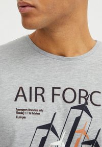 Camiseta de algodón gris con el texto en negrita "AIR FORCE" en marrón oscuro y naranja, acompañada de un diseño gráfico de formas abstractas de aviones y detalles de vuelo.
