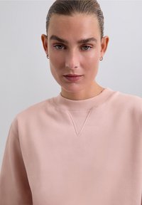 Kvinna med slickat hår som bär små hoopörhängen och en ljusrosa tröja med rund halsringning, som tittar direkt in i kameran.