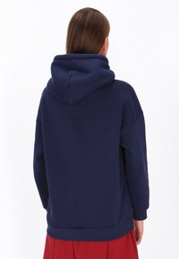 Tengerészkék kapucnis pulóver sima tapintású anyagból, nagy kapucnival és laza szabással, hátulnézetben.