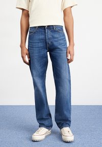 Blue denim jeans with a straight fit, featuring a classic five-pocket design and blue stitching. Worn with white sneakers.