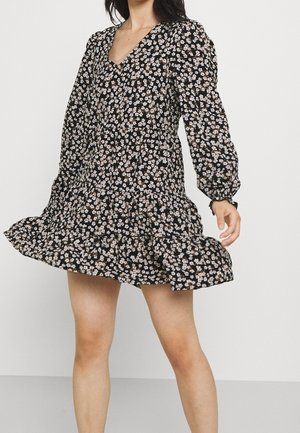 Frau trägt ein schwarzes, florales Langarmkleid mit kleinen weißen und rosa Blumen und steht vor einem schlichten grauen Hintergrund.