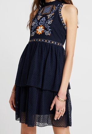 Granatowa sukienka bez rękawów z wielowarstwową spódnicą i kolorowym kwiatowym haftem na biuście, noszona przez stojącą osobę z biżuterią na dłoniach.