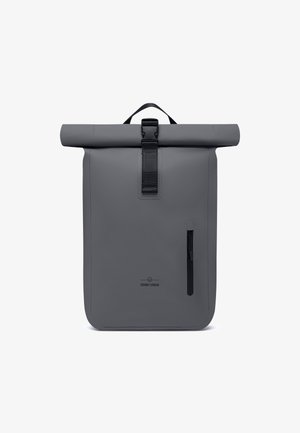 Sac à dos gris à rabat fabriqué en matériau imperméable, avec une poche zippée à l'avant et une poignée supérieure pour un transport facile. Présente un branding minimal.