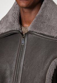 Grå shearlingjakke med en blød krave, læderfront og subtil kornstruktur; har en sølv lynlåslukning og strikket tan detalje under.