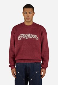 Burgundy sweater with fuzzy texture and ribbed cuffs, featuring "Pegador" logo in fluffy white lettering across the chest.
