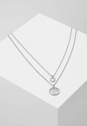 To lagdelte sølvkæder ligger på en hvid geometrisk sokkel med små runde vedhæng, hvoraf det ene er solidt og det andet har et blomsterudskåret mønster.